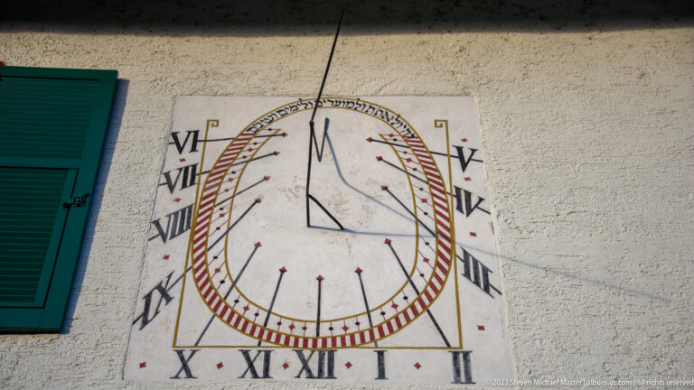 Painted oval wall sundial with Roman numerals, red hour marks, and a projecting metal gnomon beside a green shutter.