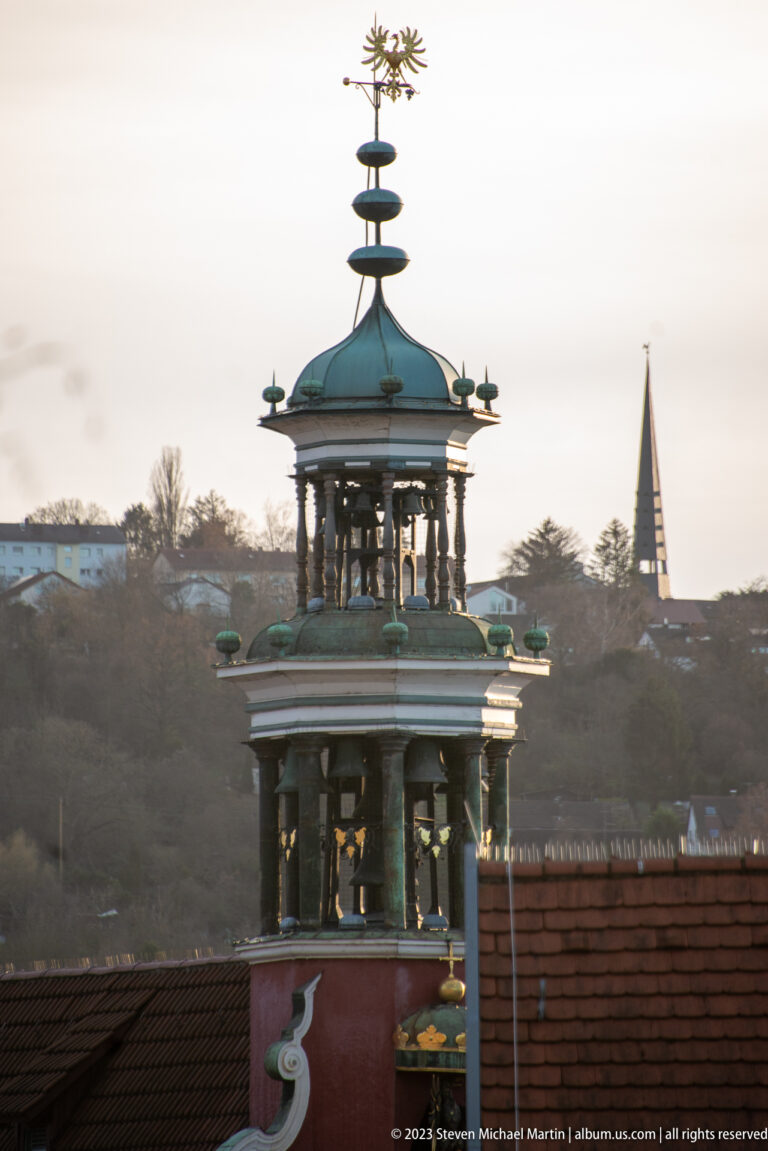 Open-sided green-patinated rooftop cupola with slender supports, stacked finial, and small projecting ornaments above red tiled roofs.