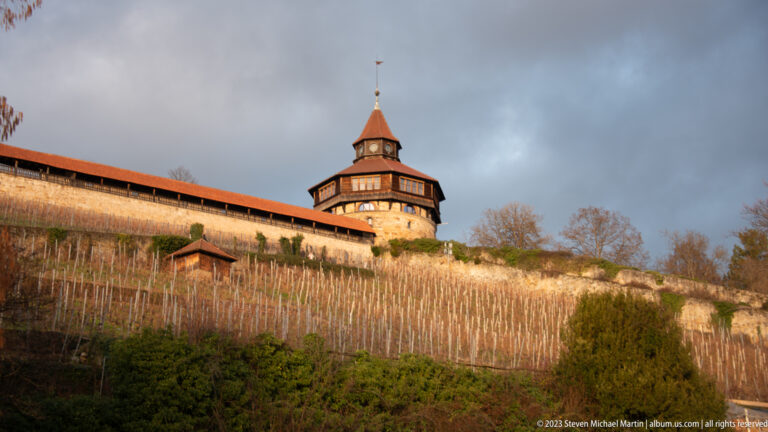 Round tower and red-roofed covered wall of Esslinger Burg above rows of winter-bare vineyard stakes.