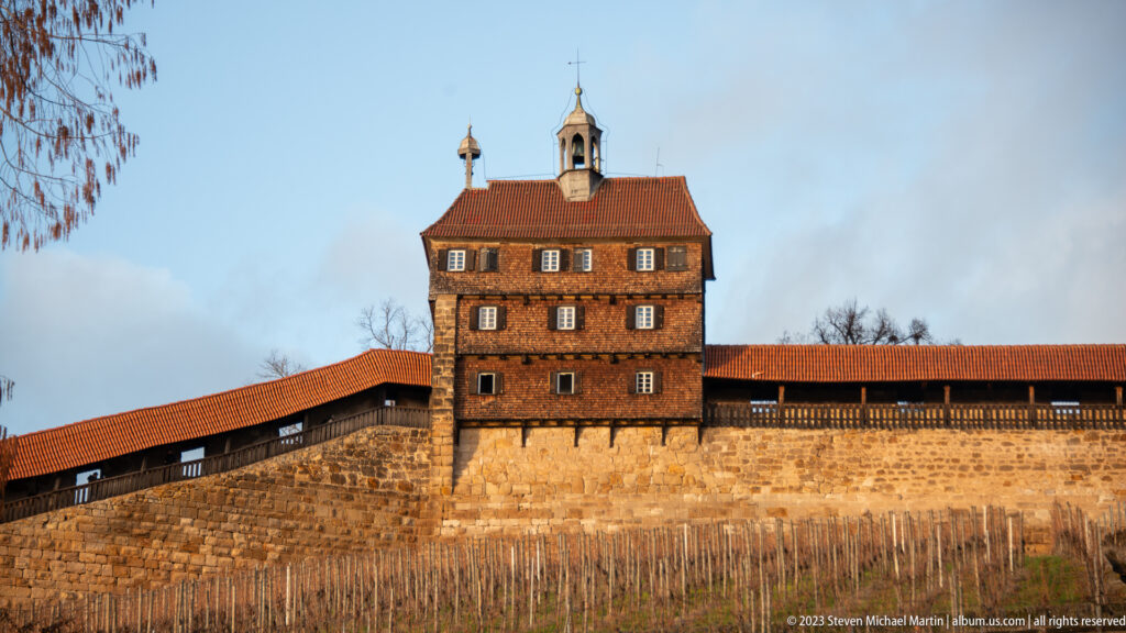 Square Esslinger Burg tower with timbered upper story and red roof rising above a stone wall and bare vineyard.