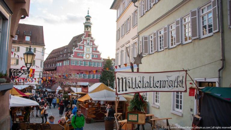 Crowded Mittelaltermarkt lane with timber stalls and overhead sign, leading toward the red-gabled Altes Rathaus.
