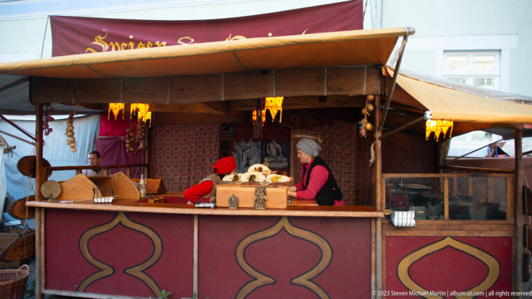 Costumed woman behind a red timber market counter with hanging goods, textiles, baskets, and warm lamps.