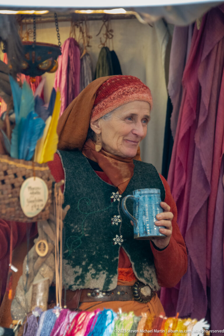 Older woman in period-inspired clothing and headscarf holding a blue mug inside a colorful medieval market stall.