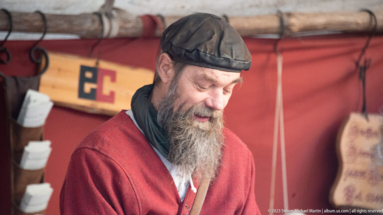 Bearded man in dark cap and red tunic looking downward inside a medieval-themed market stall.