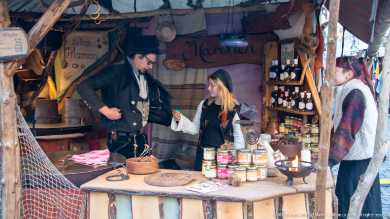 Three costumed vendors inside a rustic timber stall stocked with jars, bottles, baskets, bowls, and hanging signs.