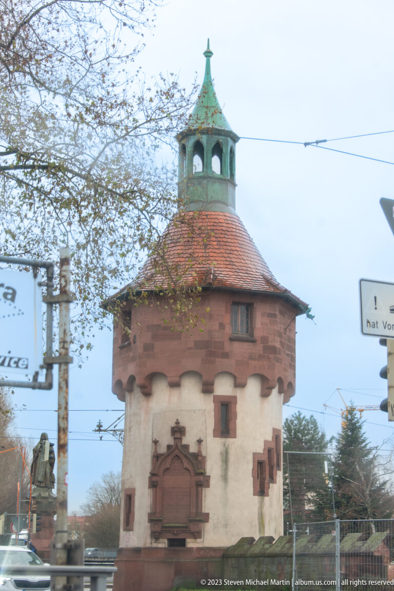 Round pale masonry tower with tiled polygonal roof and green open lantern beside a Freiburg street.