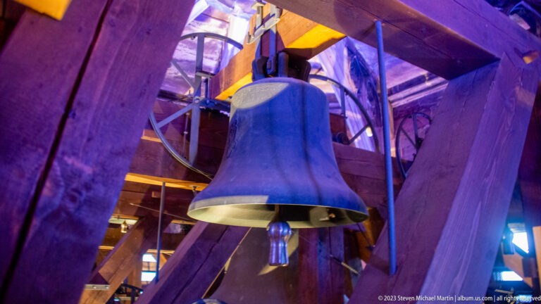 Bronze bell viewed from below among heavy timber beams and metal fittings in Freiburg Minster belfry.