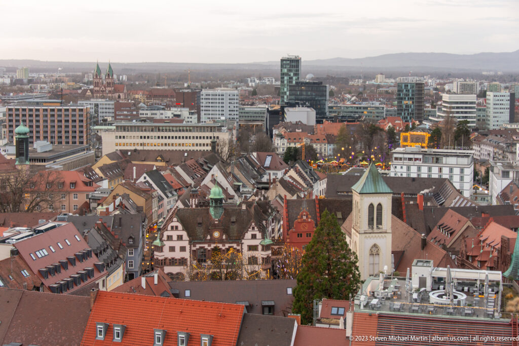 Elevated view over Freiburg red-tiled roofs, church towers, modern buildings, and distant low hills.