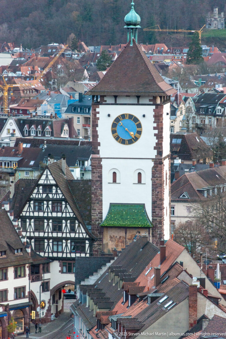 White historic gate tower with blue-and-gold clock, steep tiled roof, and green lantern above Freiburg rooftops.
