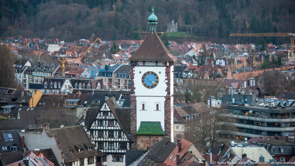 Clock-faced historic gate tower rising above Freiburg rooftops with wooded hills in the background.