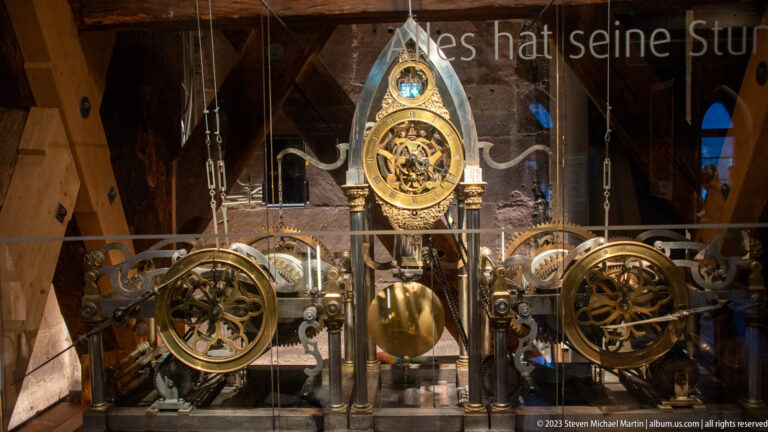 Historic clock mechanism behind glass with large brass gears, pendulum, dials, and pointed central frame.