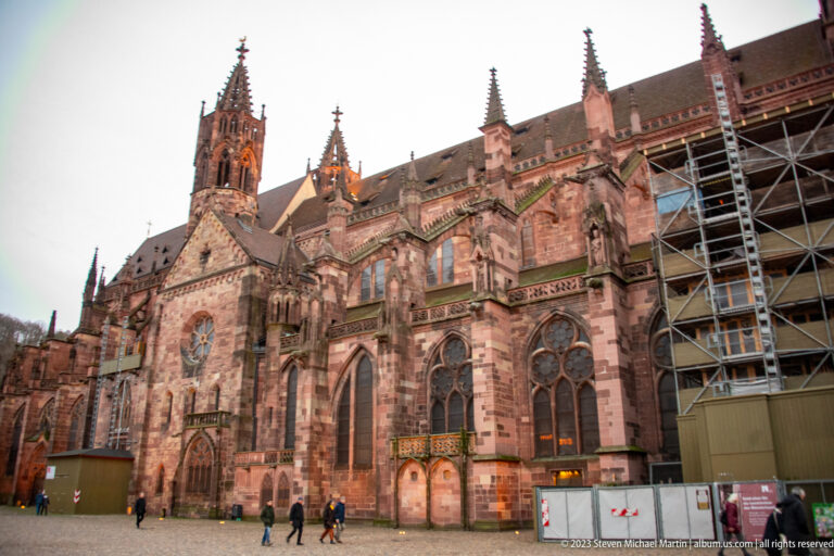 Side exterior of Freiburg Minster with red sandstone Gothic windows, buttresses, pinnacles, tower, and scaffolding.