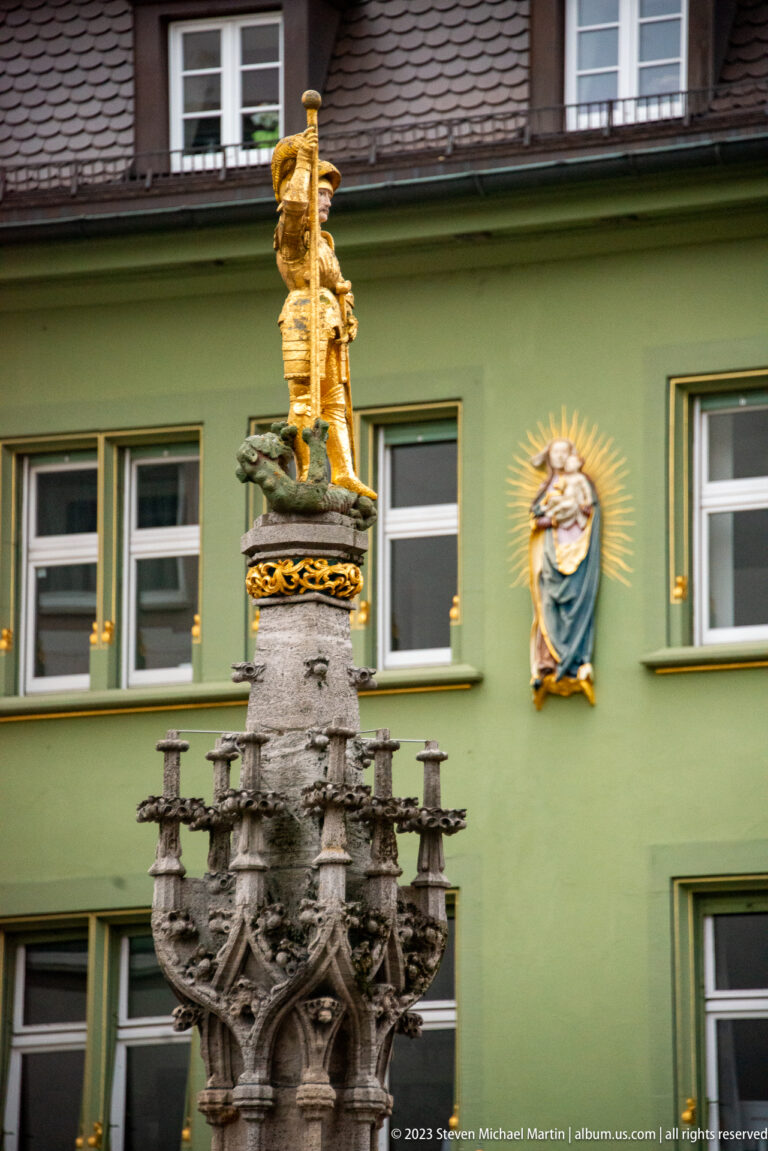 Gilded figure crowning an ornate stone fountain against a green building with a radiant wall emblem.