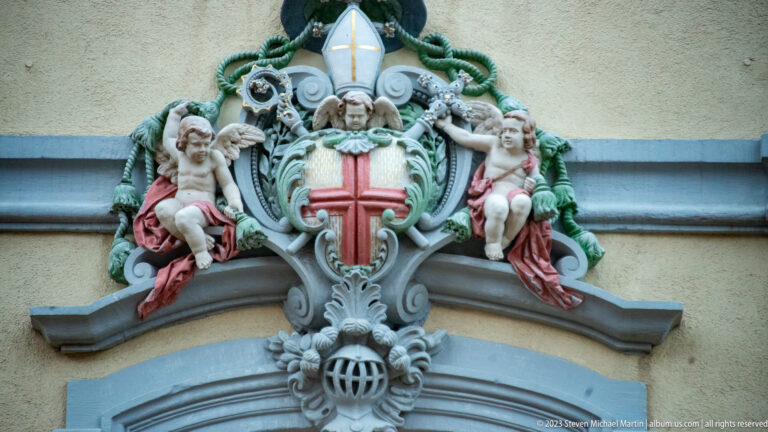 Ornate coat of arms above a doorway with red cross, bishop’s mitre, cords, scrollwork, and two cherubs.