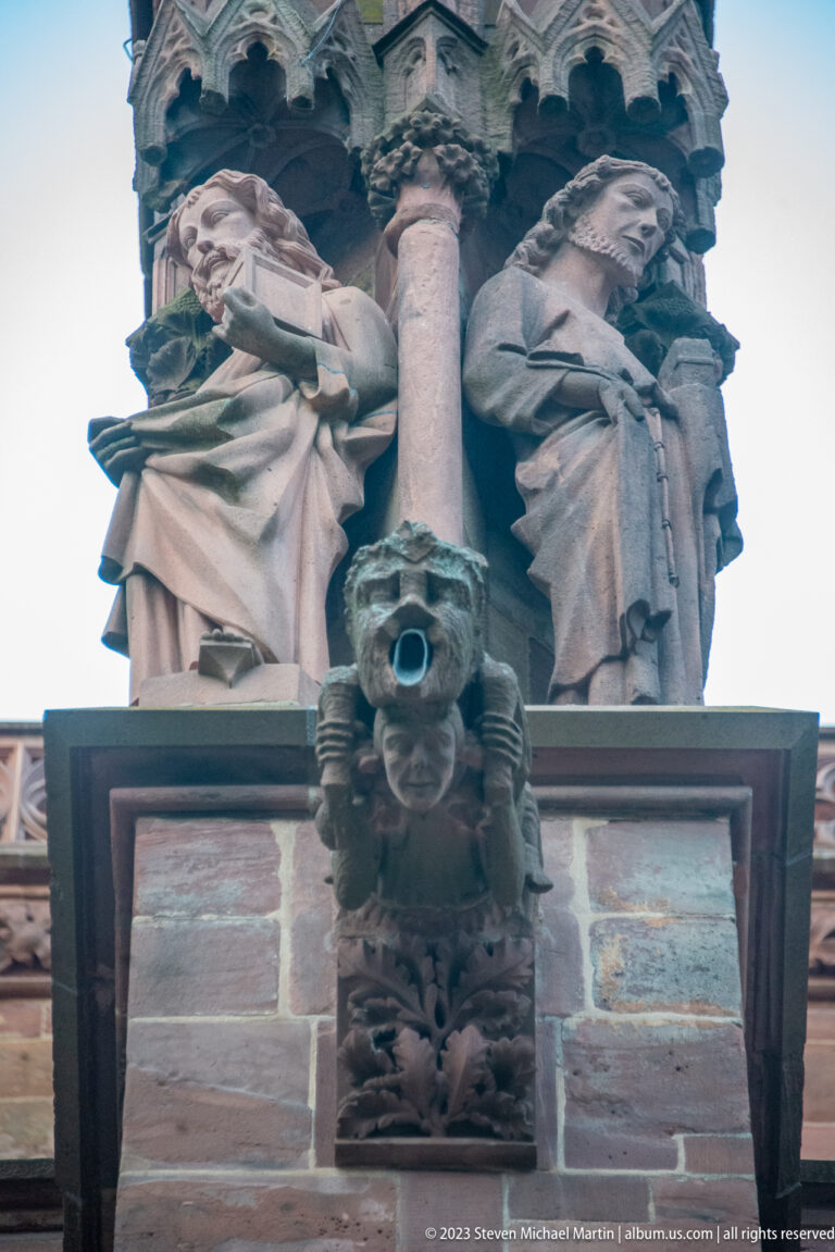 Two weathered seated stone figures flanking a shaft above a carved projection on Freiburg Minster exterior.
