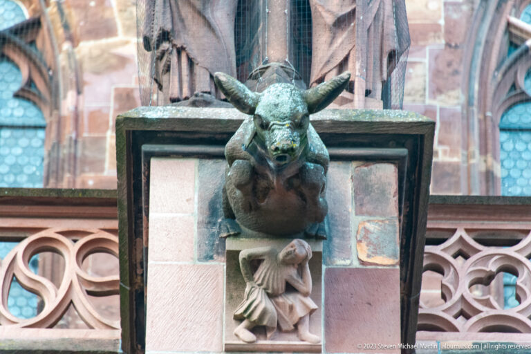 Horned animal-headed gargoyle with open mouth projecting above a smaller carved figure on Freiburg Minster.