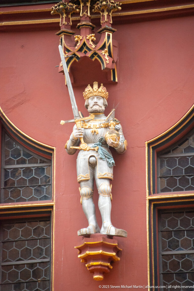 Polychrome armored ruler holding a sword on a gilded bracket against the red Historisches Kaufhaus façade.