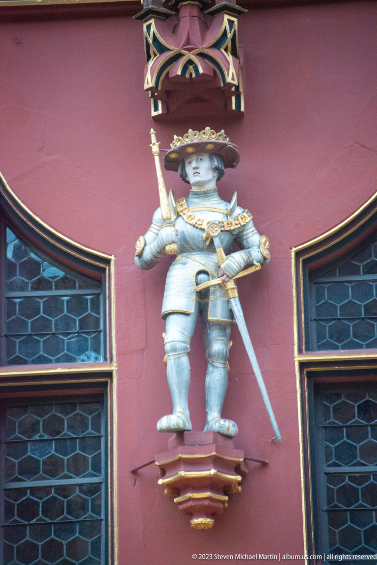 Armored ruler statue with long sword between leaded windows on the red façade of Freiburg Historisches Kaufhaus.