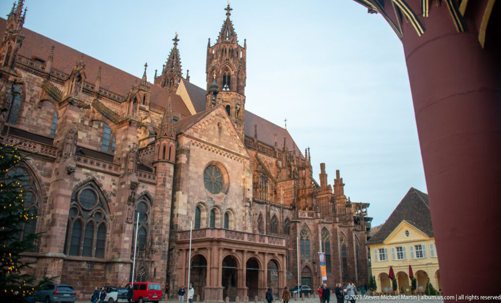 Freiburg Minster seen across Münsterplatz through a red arcade arch of the Historisches Kaufhaus.