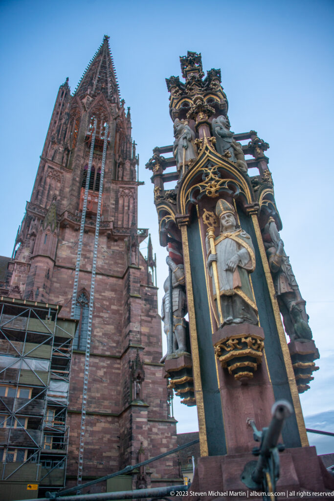 Gilded fountain figure beneath an ornate canopy with Freiburg Minster tower rising in the background.