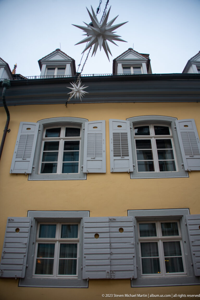 Large many-pointed illuminated star suspended above a yellow Freiburg façade with white shutters and dormer windows.