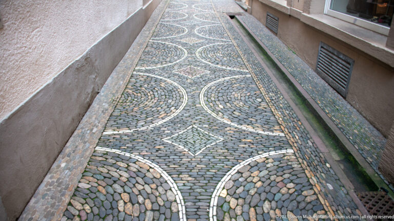 Ornamental pebble paving with circular patterns and a central diamond fills a narrow passage between Freiburg buildings.