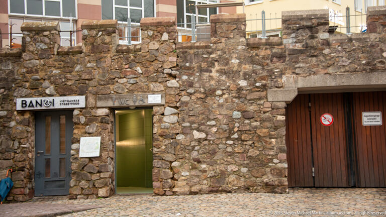Rough historic stone-and-brick wall with three modern doorways and small signs along a cobbled Freiburg street.