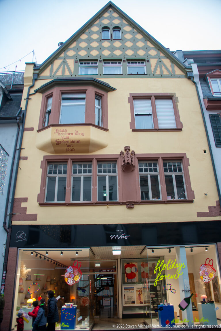 Narrow yellow historic façade with red trim, projecting bay, patterned gable, and brightly lit shop windows at street level.