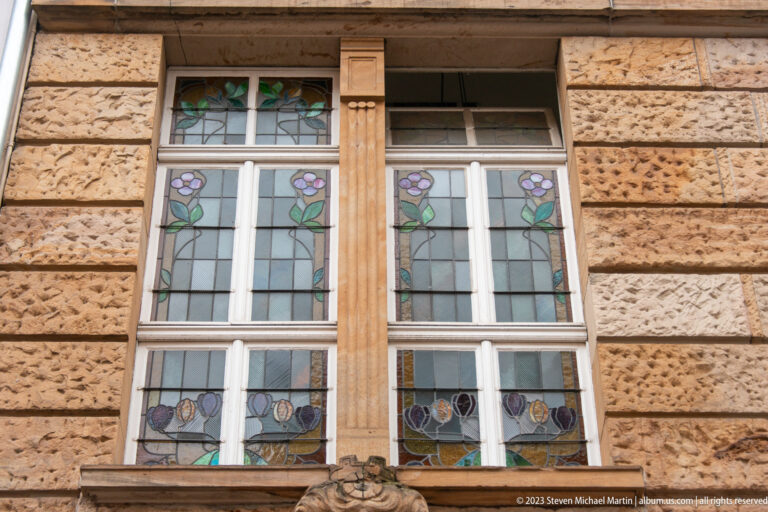 Paired tall windows with floral stained glass set between carved mullions in a heavily rusticated stone façade.