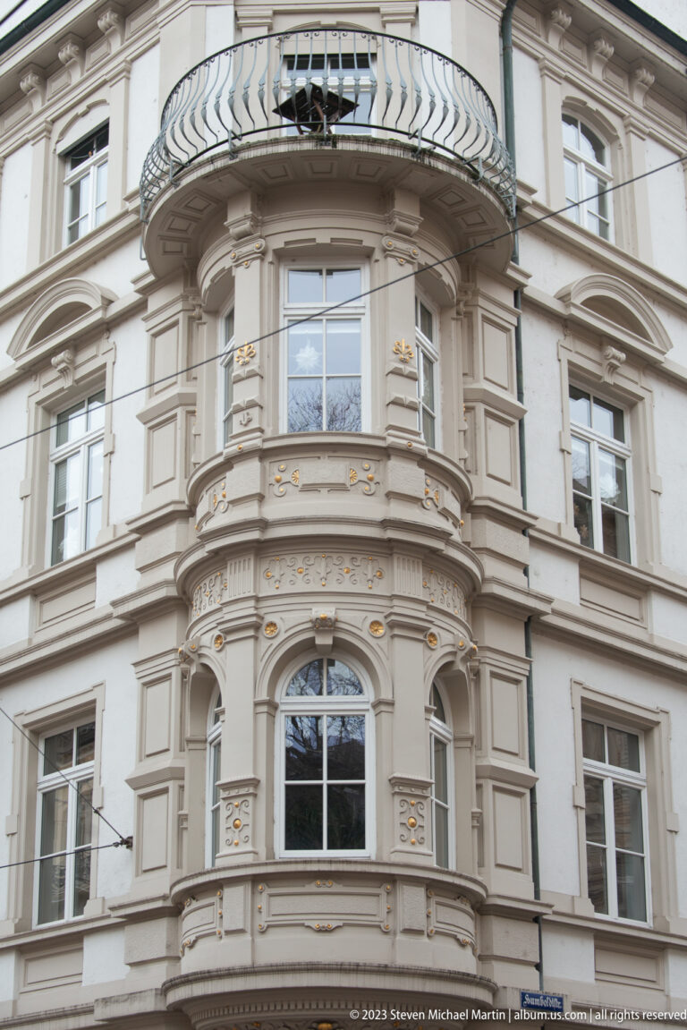 Ornate pale corner façade with rounded bay windows, sculpted moldings, gold accents, and a curved wrought-metal balcony.
