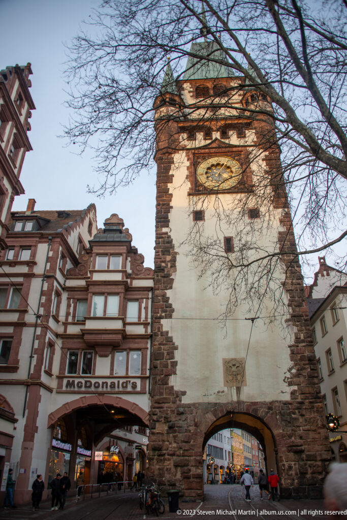 Schwabentor city gate with arched passage, white tower, stone corners, large clock face, and shops beside it in Freiburg.