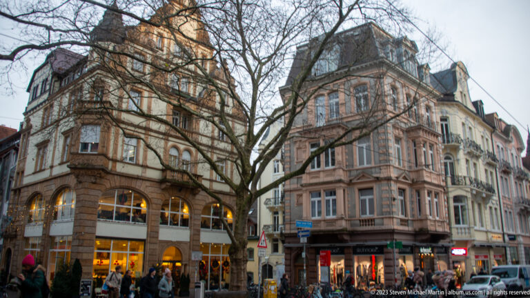 Two ornate multi-story corner buildings with bay windows and shops frame a busy Freiburg intersection beneath leafless trees.