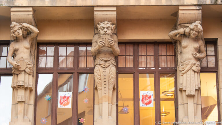 Three carved stone human figures stand between large windows, with Salvation Army emblems visible inside the Freiburg building.
