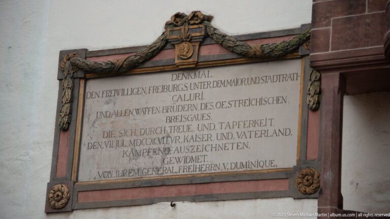German-language memorial plaque in a carved wreath-like frame mounted on a Freiburg wall beside reddish stone trim.