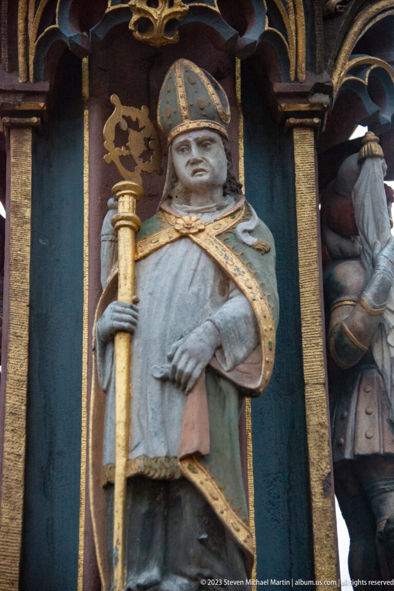 Carved bishop or saint in mitre and cope holding a staff within a gilded fountain niche.