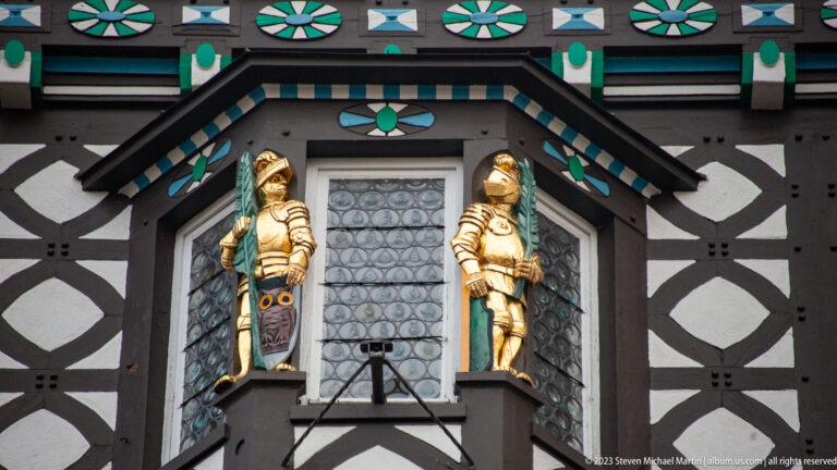 Two gilded armored figures flank a small leaded window on a black, white, turquoise, and green half-timbered façade.