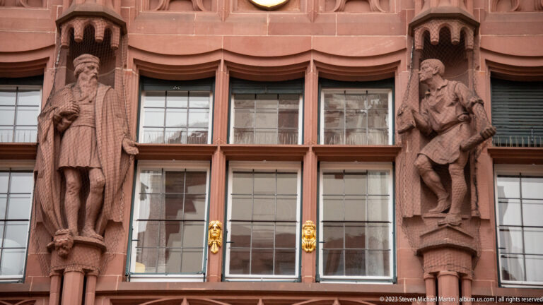 Two carved male figures in historical dress stand between windows on a reddish stone façade with small gilded ornaments below.
