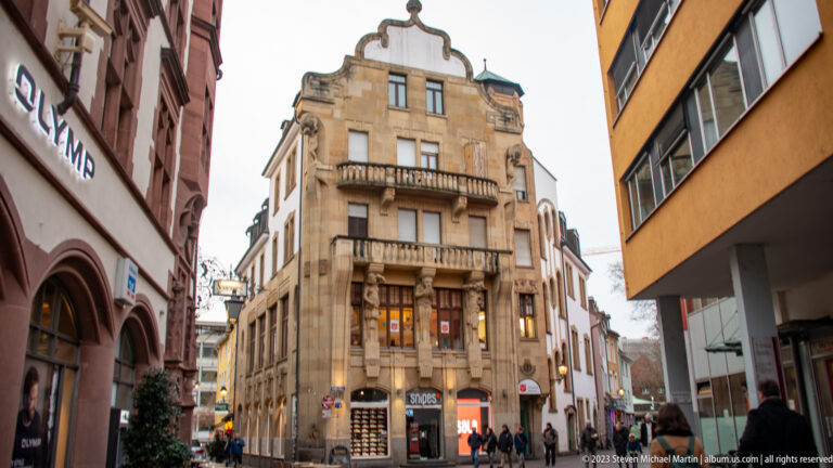 Tall pale stone corner building with balconies and a curved gable rises above shops and pedestrians on a narrow Freiburg street.