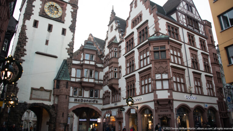 Gabled historic buildings with projecting bays and shops stand beside the arched Martinstor passage in Freiburg’s old town.