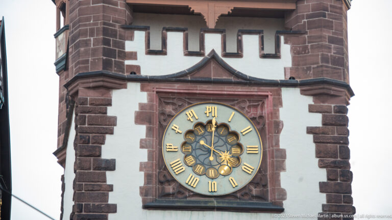 Black-and-gold Roman-numeral clock set in a red-stone frame on the white upper façade of Martinstor.