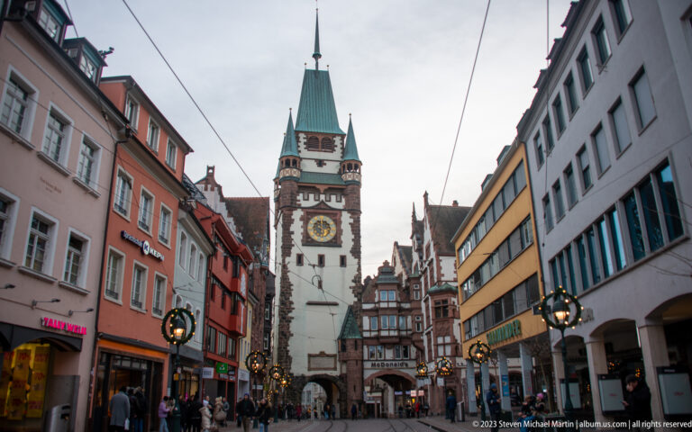 Martinstor clock tower rises over its pointed gateway at the end of a pedestrian shopping street with tram wires and holiday decorations.