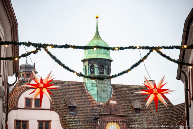 Two glowing red star lanterns and evergreen garlands frame a green copper turret and tiled historic roof in Freiburg.