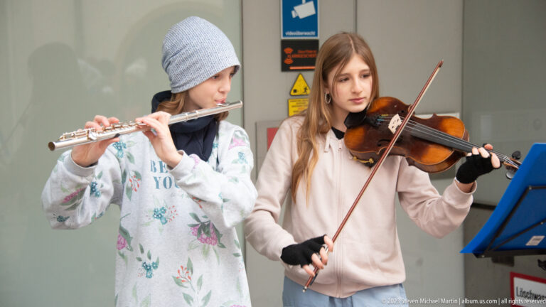 Two young street musicians, one playing flute and one violin, perform beside a blue music stand in Freiburg.