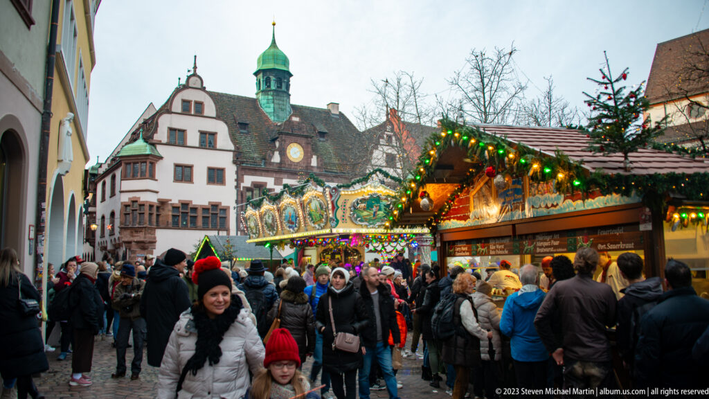 Crowded Freiburg Christmas market with lit wooden stalls, evergreen decorations, a colorful attraction, and historic buildings behind.