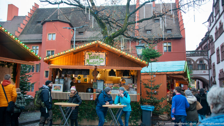 Visitors eat and drink at standing tables outside a lit wooden Christmas-market stall in front of a red historic building.