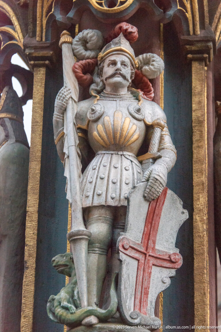 Armored carved figure holding a sword beside a white shield marked with a red cross.