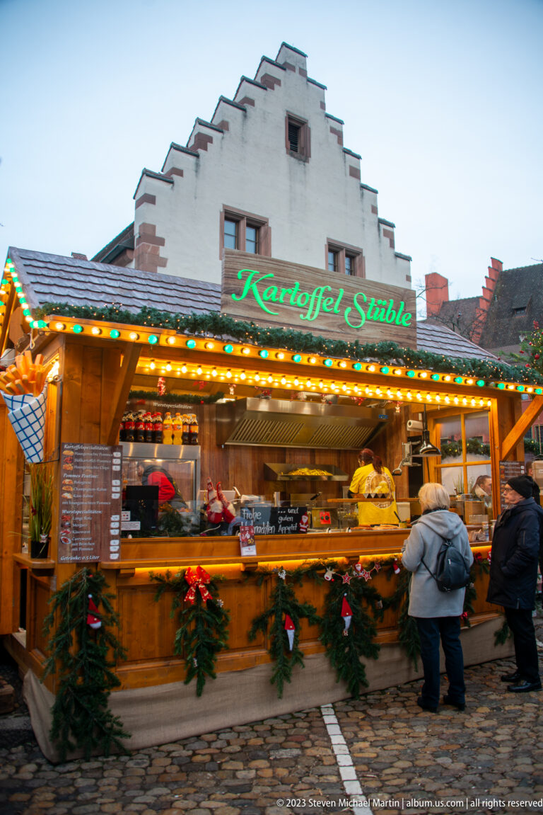 Illuminated Kartoffel Stüble food stall with customers, evergreen trim, and a stepped-gable building behind in Freiburg.