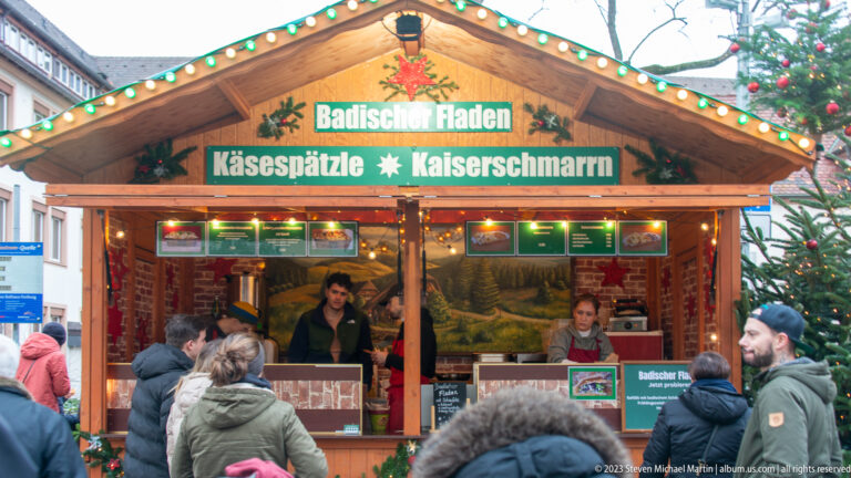 Wooden Freiburg market stall labeled Badische Fladen, Käsespätzle and Kaiserschmarrn, with customers gathered at the counter.