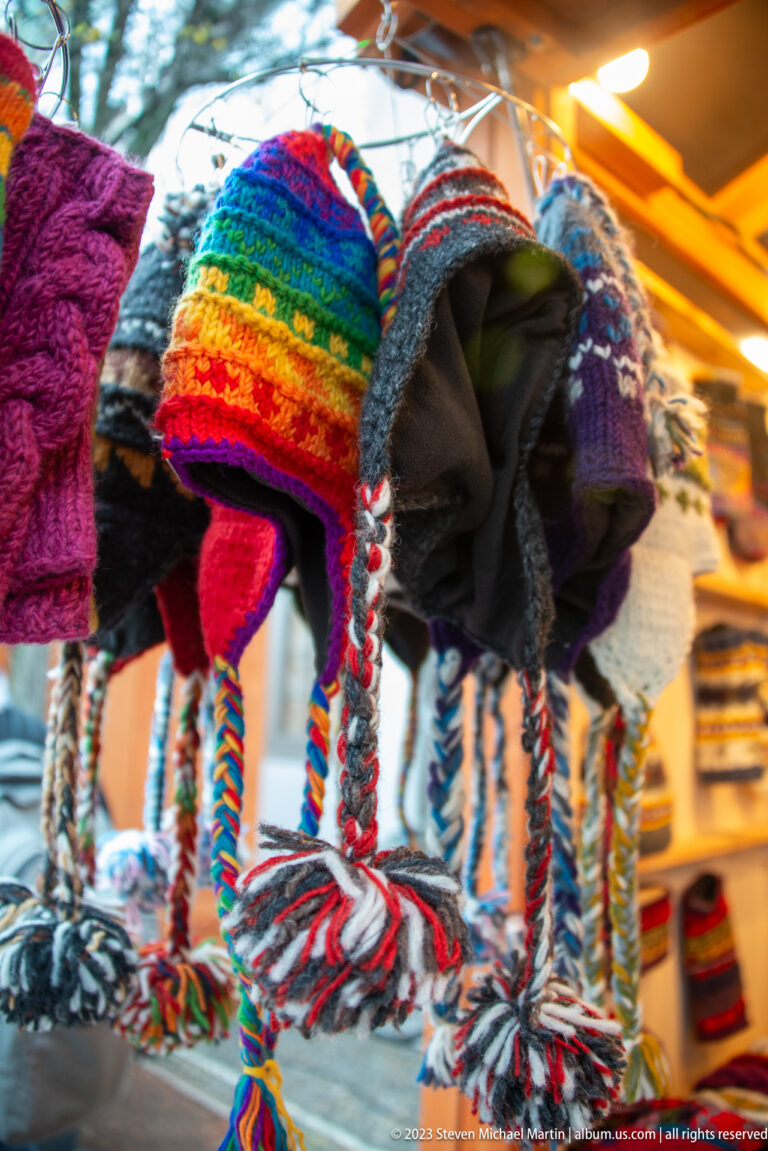 Rows of colorful knitted winter hats with stripes, earflaps, braids, and pom-poms hang at a Freiburg market stall.