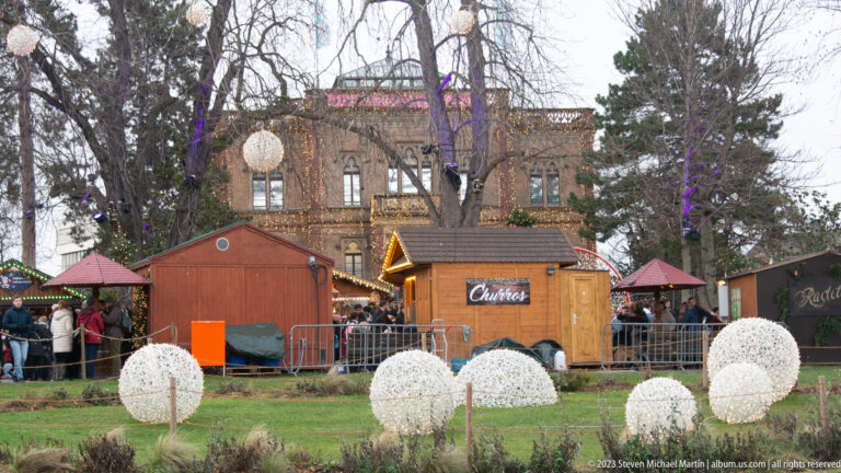 Wooden Christmas-market huts and glowing white spheres stand on the lawn before the ornate Colombischlössle in Freiburg.